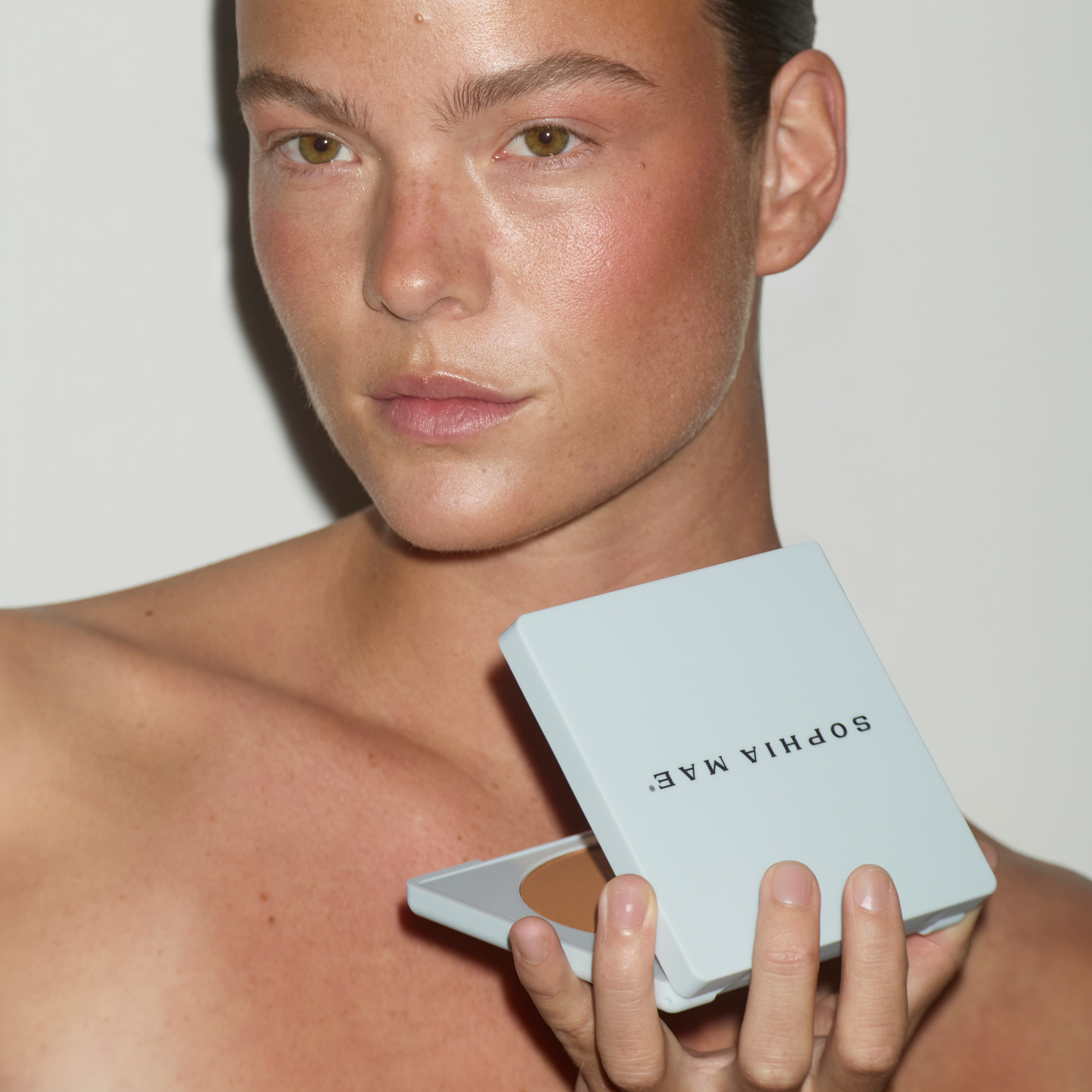 Model holding an open SOPHIA MAE Matte Bronzer compact while wearing the bronzer on her face.