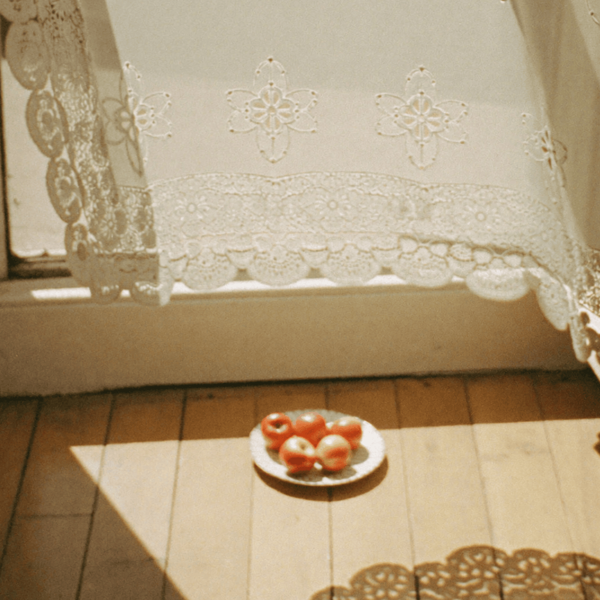 Sunlit kitchen table with a plate of red apples beneath embroidered lace curtains, creating a warm and nostalgic atmosphere.