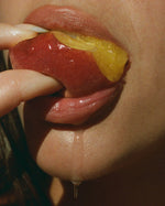 Close-up of glossy lips biting a juicy peach, evoking the fruity feel of Soft Summer Kiss EDP by SOPHIA MAE.