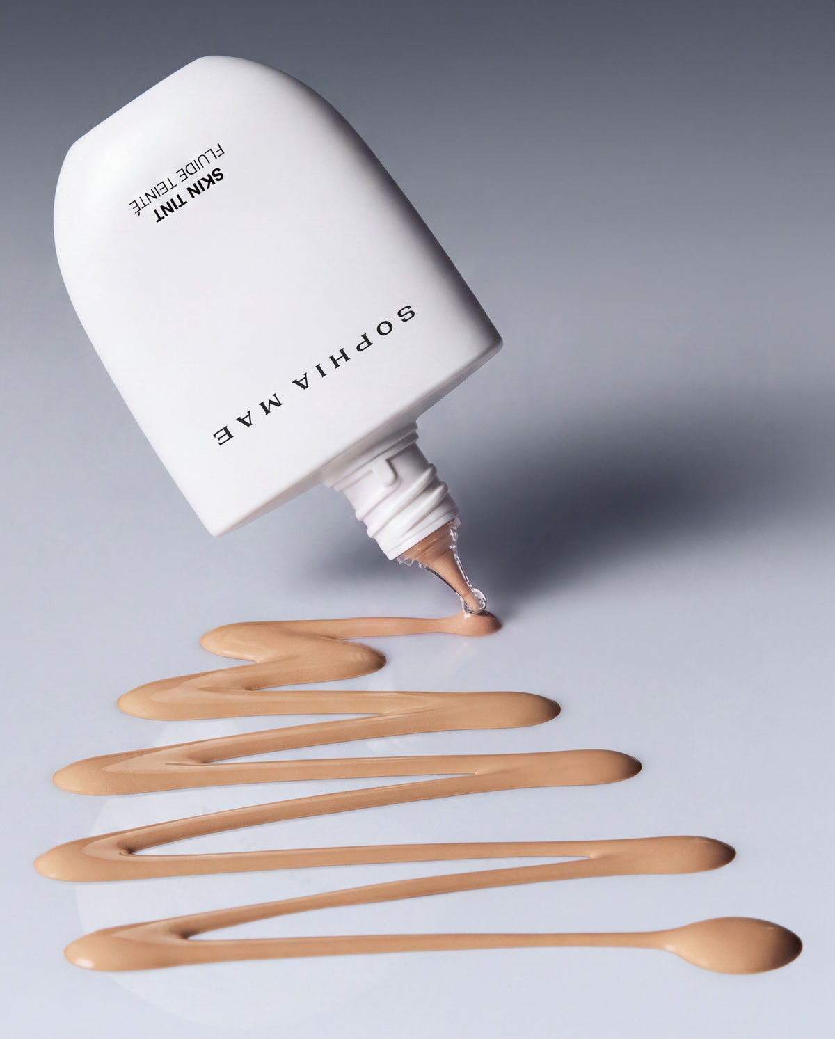 SOPHIA MAE Skin Tint dispensing onto a neutral surface, highlighting its fluid texture and lightweight, blendable formula.