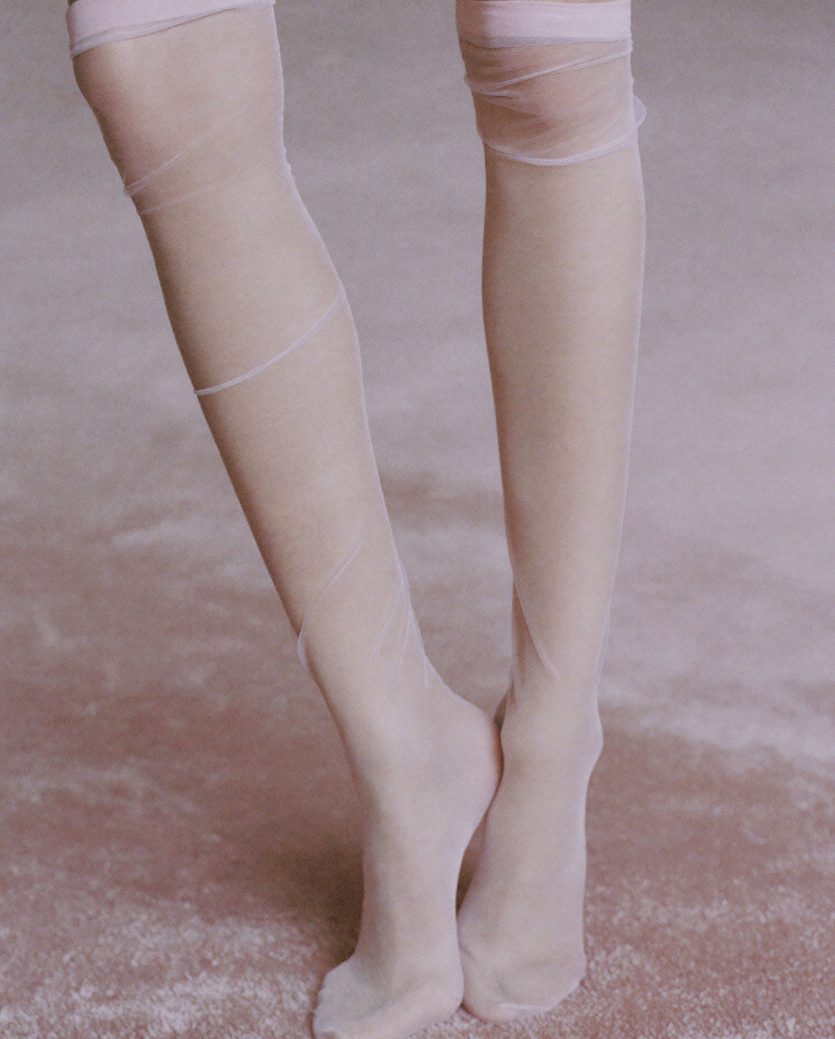 Close-up of legs in sheer ivory tights on a soft pink carpet, reflecting the delicate aesthetic of Whispers of Elegance eau de parfum.