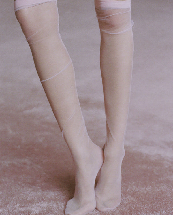 Close-up of legs in sheer ivory tights on a soft pink carpet, reflecting the delicate aesthetic of Whispers of Elegance eau de parfum.