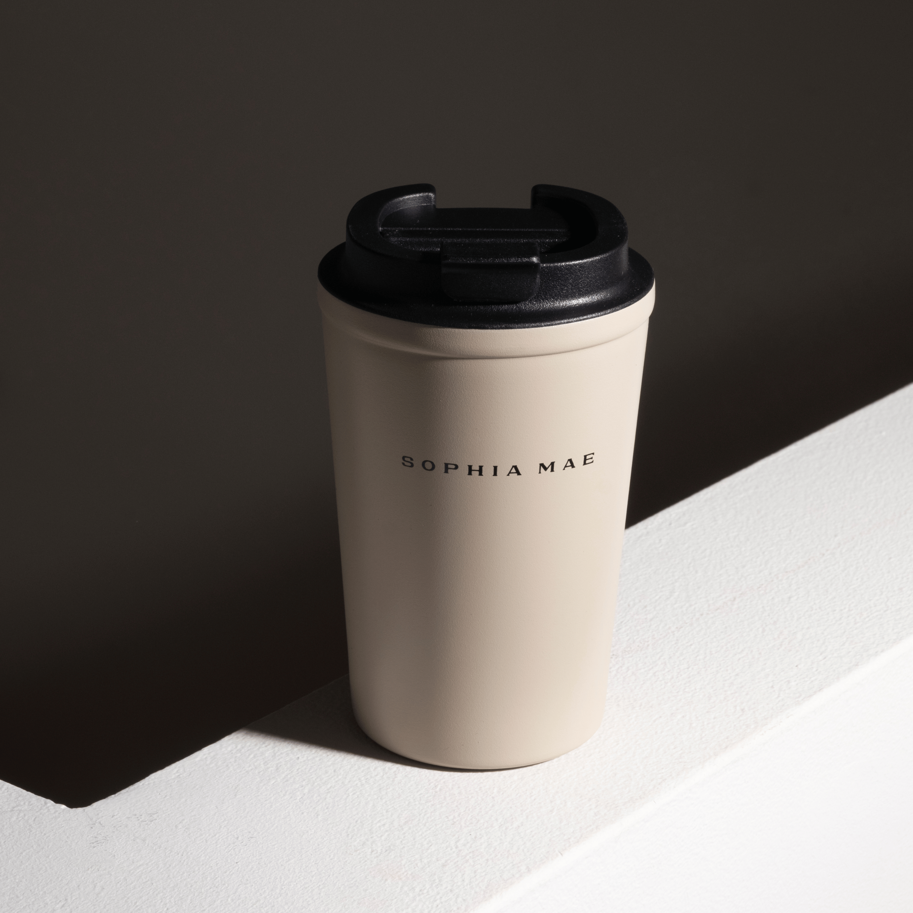 Beige SOPHIA MAE Travel Mug with black lid in a minimalist sunlit setting.