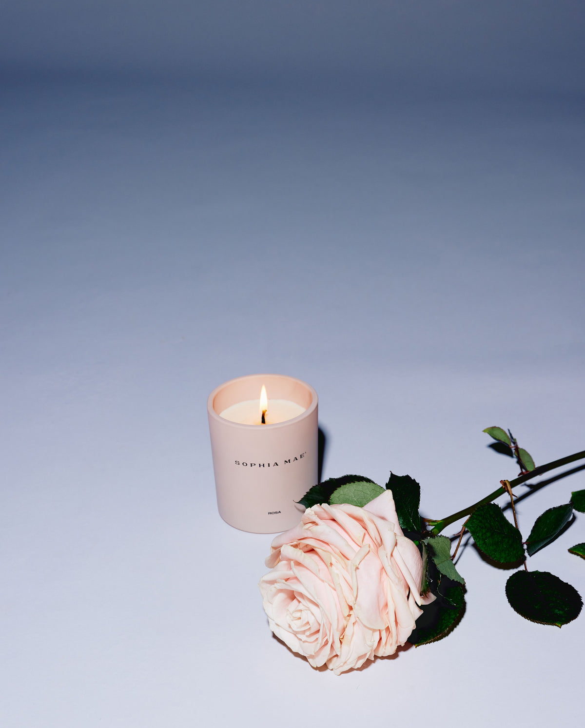 SOPHIA MAE Rosa scented candle burning beside a pink rose on a soft blue background.