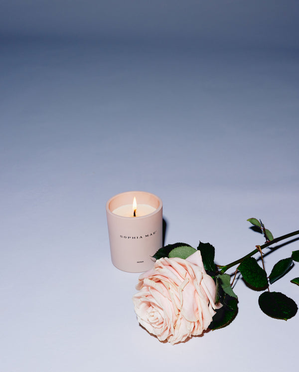 SOPHIA MAE Rosa scented candle burning beside a pink rose on a soft blue background.