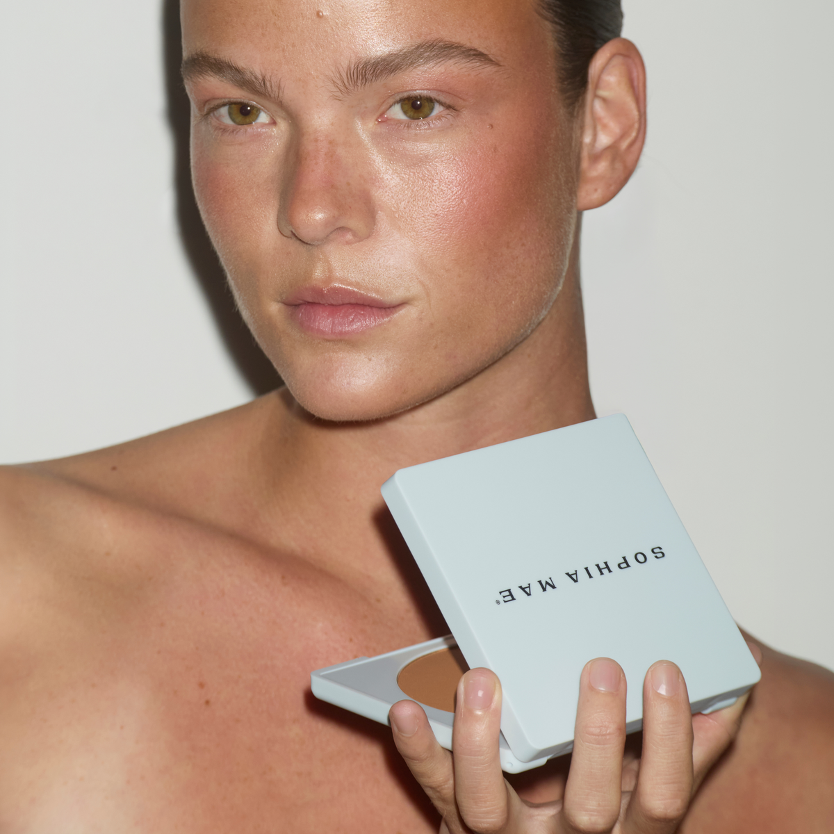 Model holding an open SOPHIA MAE Matte Bronzer compact while wearing the bronzer on her face.