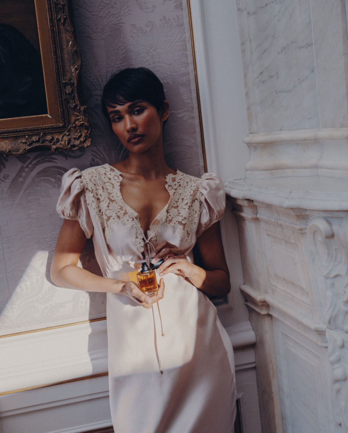 Model in a satin lace dress holding SOPHIA MAE Still in Bloom Eau de Parfum in an elegant interior.