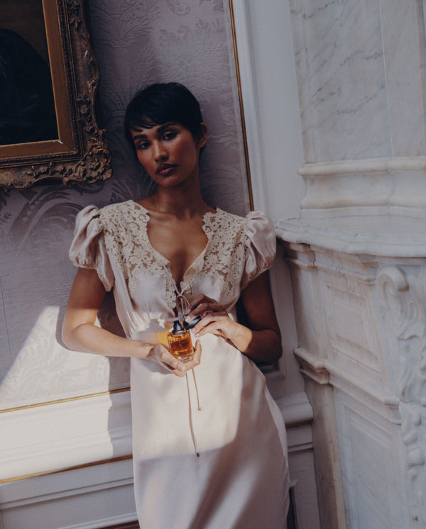 Model in a satin lace dress holding SOPHIA MAE Still in Bloom Eau de Parfum in an elegant interior.