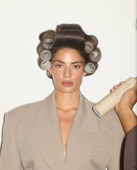 Model wearing SOPHIA MAE Velcro Hair Rollers while a volumizing hair spray is applied, capturing the styling process for lifted, full-bodied hair.