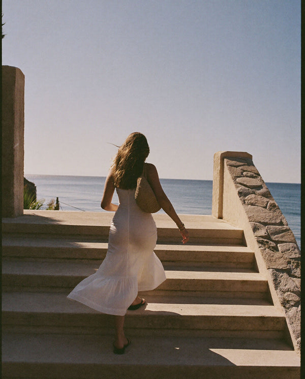 Woman in a flowing white dress walking toward the sea, inspired by the warm coastal feeling of Golden Mirage perfume by SOPHIA MAE.