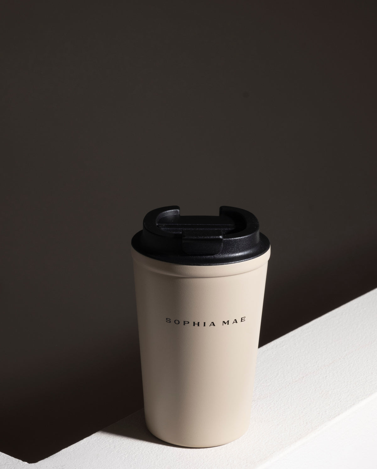SOPHIA MAE Travel Mug in cream with black lid, styled on a minimal neutral surface.