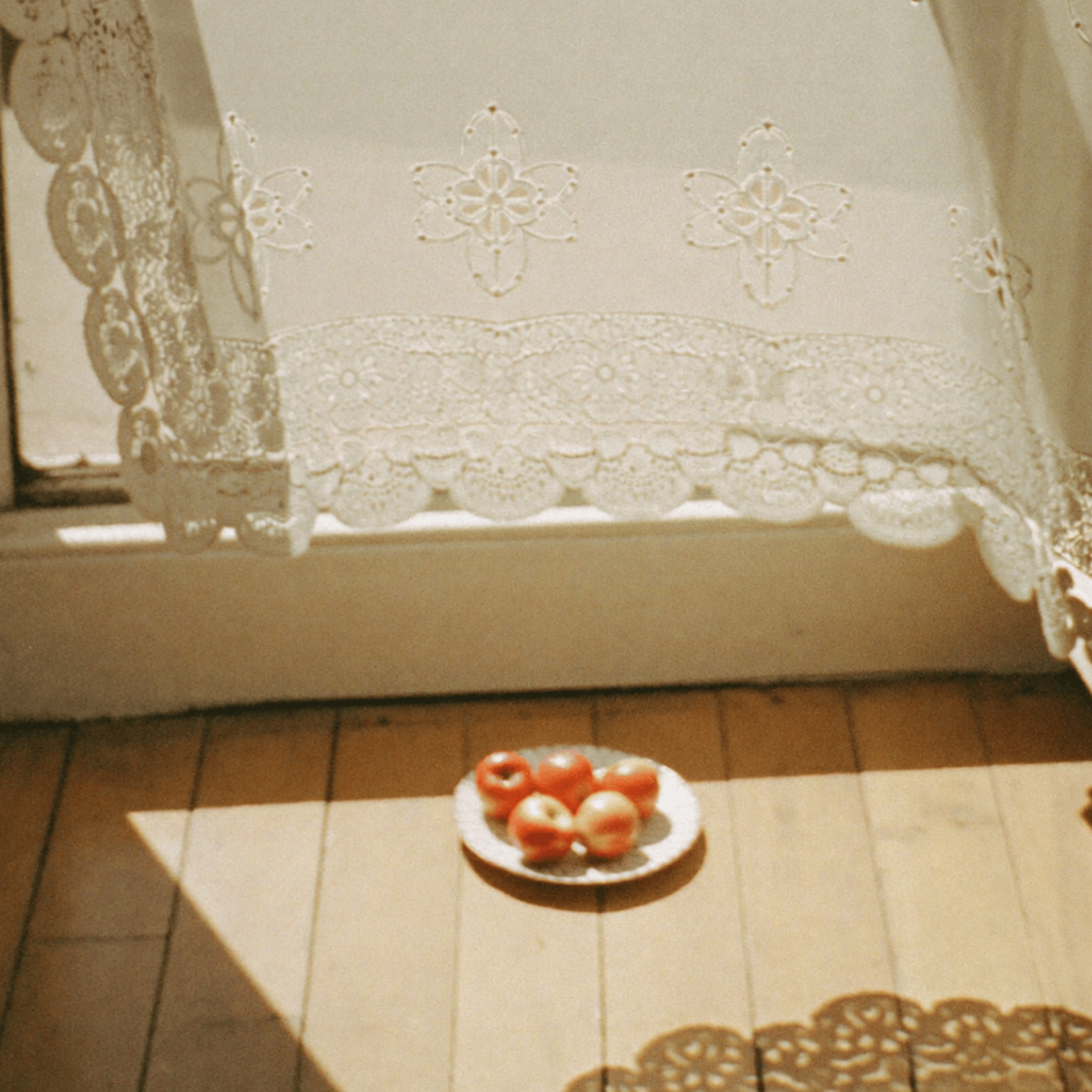 Sunlit kitchen table with a plate of red apples beneath embroidered lace curtains, creating a warm and nostalgic atmosphere.