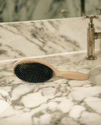SOPHIA MAE Hairbrush resting on a marble vanity, featuring dense black boaar bristles and a smooth beige handle in a refined bathroom setting.