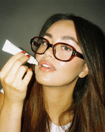 Model applying SOPHIA MAE Liquid Lip Balm while wearing glasses.