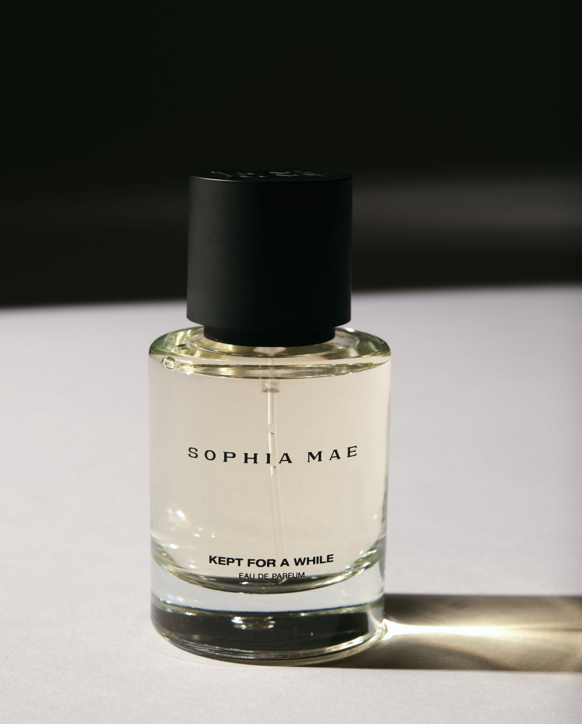 Close-up of SOPHIA MAE Kept for a While eau de parfum in a clear glass bottle, illuminated by warm sunlight against a dark background.