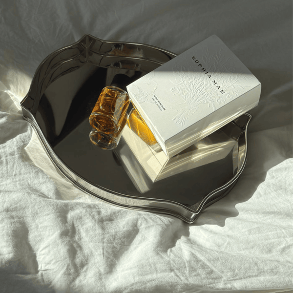 SOPHIA MAE Still in Bloom eau de parfum and its embossed white packaging arranged on a reflective silver tray in warm sunlight.