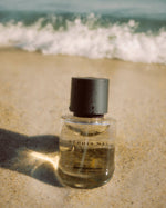 SOPHIA MAE Written in Sand Eau de Parfum bottle resting on the sand with ocean waves in the background.